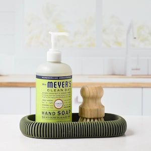 May include: A green, ribbed soap dish holds a bottle of Mrs. Meyer's Clean Day Lemon Verbena hand soap and a wooden handled brush. The soap bottle has a white pump and a green and yellow label. The brush has light-colored bristles.