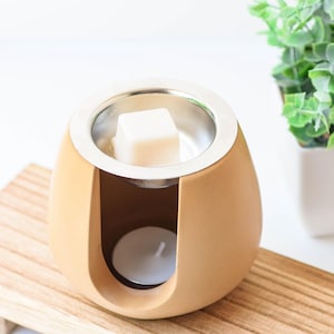 May include: A tan ceramic wax warmer with a silver dish and a white wax melt. A small white tea light candle is inside the warmer. The warmer sits on a wooden tray, with a small green plant in the background.