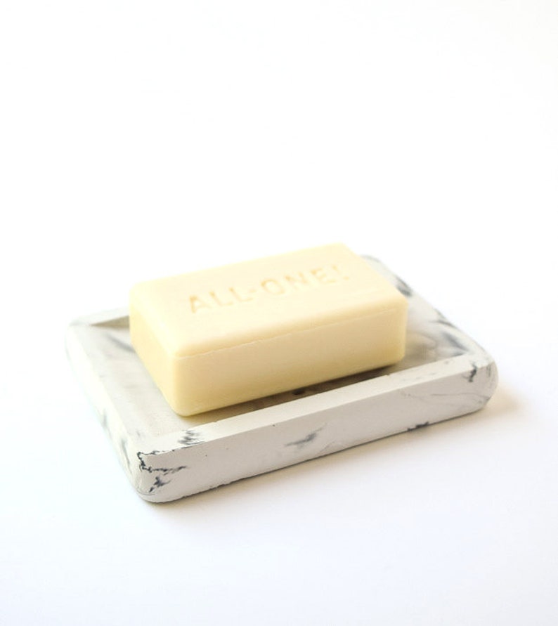 May include: A white soap dish with a marble pattern and a bar of soap that says "ALL ONE" on it.