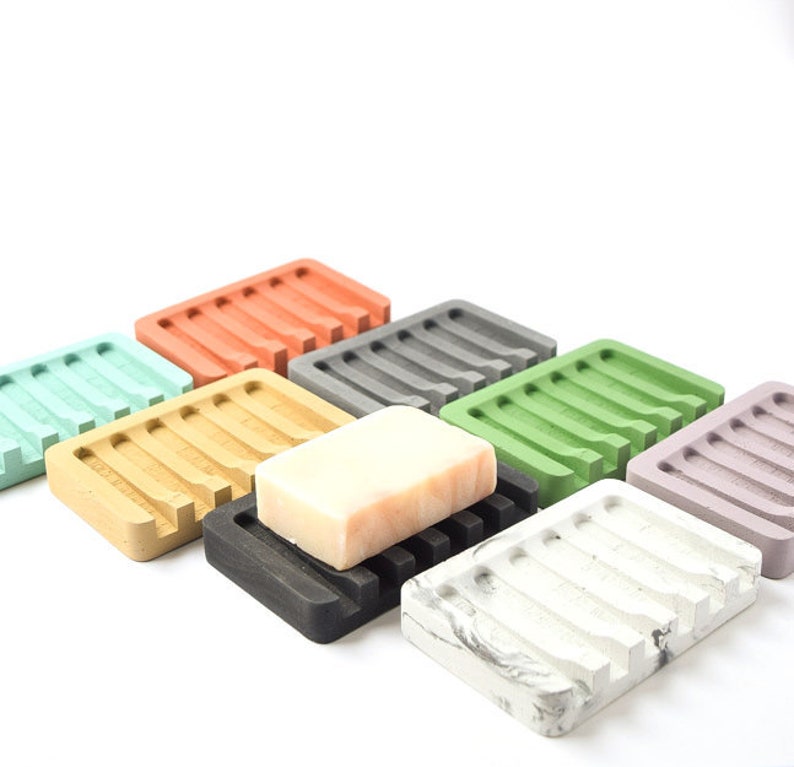 May include: Assortment of colorful soap dishes made of concrete, with a bar of soap on one of the dishes. The dishes are in various colors, including green, gray, orange, beige, and white.