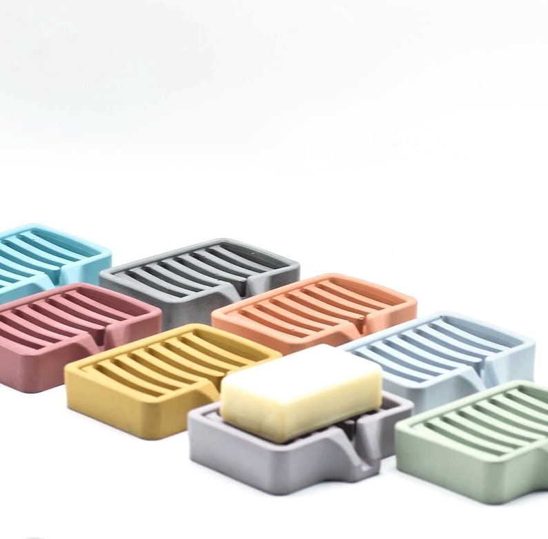 K&ouml;nnte beinhalten: Eine Sammlung farbenfroher Seifenschalen in verschiedenen Farbt&ouml;nen, darunter Blau, Grau, Rosa und Gelb. Jede Schale hat ein geschlitztes Design und eine kleine Lippe. Eine Schale enth&auml;lt ein St&uuml;ck Seife.