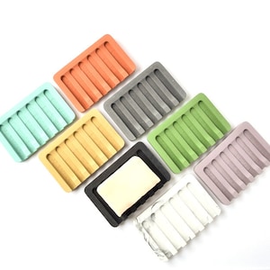 May include: Set of eight soap dishes in various colors, including light green, yellow, gray, green, purple, white, and orange. The dishes are made of concrete and have a ridged design. One dish is black and has a bar of soap inside.