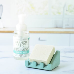 May include: A light green soap dish with a white bar of soap inside. A bottle of Branch Basics foaming wash is in the background.