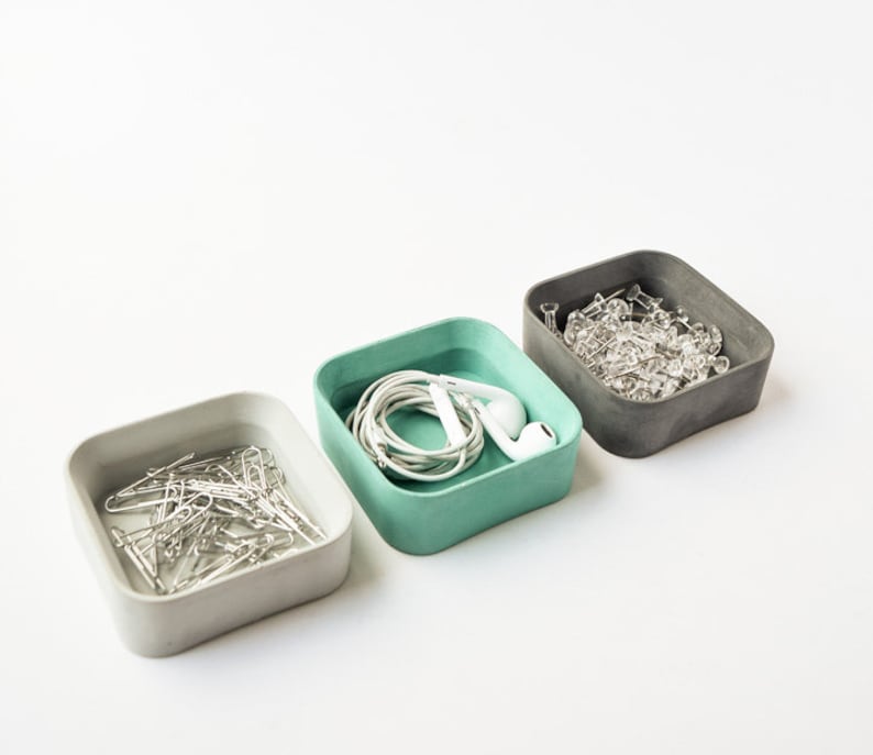 K&ouml;nnte beinhalten: Drei quadratische Betonbeh&auml;lter in Grau, Hellgr&uuml;n und Wei&szlig;, die jeweils mit verschiedenen Gegenst&auml;nden gef&uuml;llt sind. Der graue Beh&auml;lter enth&auml;lt durchsichtige Stecknadeln, der hellgr&uuml;ne Beh&auml;lter enth&auml;lt wei&szlig;e Ohrh&ouml;rer und ein wei&szlig;es Kabel, und der wei&szlig;e Beh&auml;lter enth&auml;lt silberne B&uuml;roklammern.