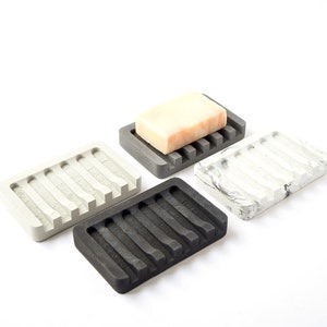 May include: Four rectangular soap dishes made of concrete in different colors: gray, black, white with gray marbling, and gray with a bar of soap on top. Each dish has a series of raised ridges on the bottom to help drain water.