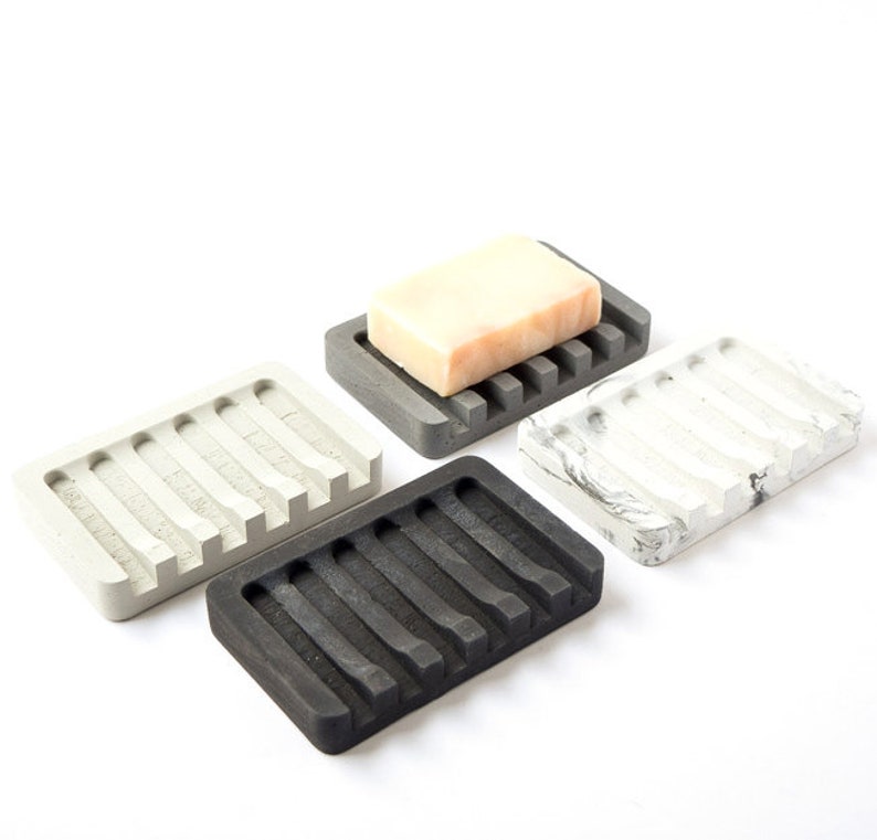 May include: Four rectangular soap dishes made of concrete in different colors: gray, black, white with gray marbling, and gray with a bar of soap on top. Each dish has a series of raised ridges on the bottom to help drain water.