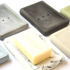 May include: A set of five rectangular soap dishes made of concrete in various colors, including gray, black, white, and green. One dish has a bar of soap on top that says "ALL ONE!" in yellow lettering.
