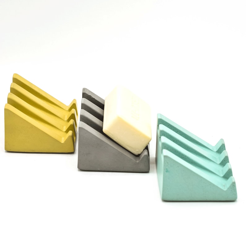 May include: Three soap dishes made of concrete in yellow, gray, and light blue. The gray dish has a bar of soap on top.