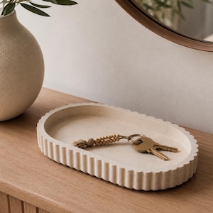 May include: An oval-shaped beige tray with a fluted, scalloped edge design. Inside the tray rests a metal keyring featuring a small tassel and a series of light-colored wooden beads attached to a circular ring with two brass-colored keys.