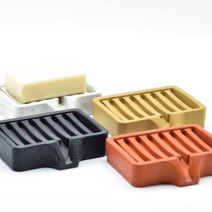 K&ouml;nnte beinhalten: Vier Seifenschalen in verschiedenen Farben: Schwarz, Wei&szlig;, Beige und Orange. Jede Schale hat eine rechteckige Form mit einem Lamellendesign und einem kleinen abgewinkelten Abschnitt zur Entw&auml;sserung. Eine Seife liegt auf der wei&szlig;en Schale.