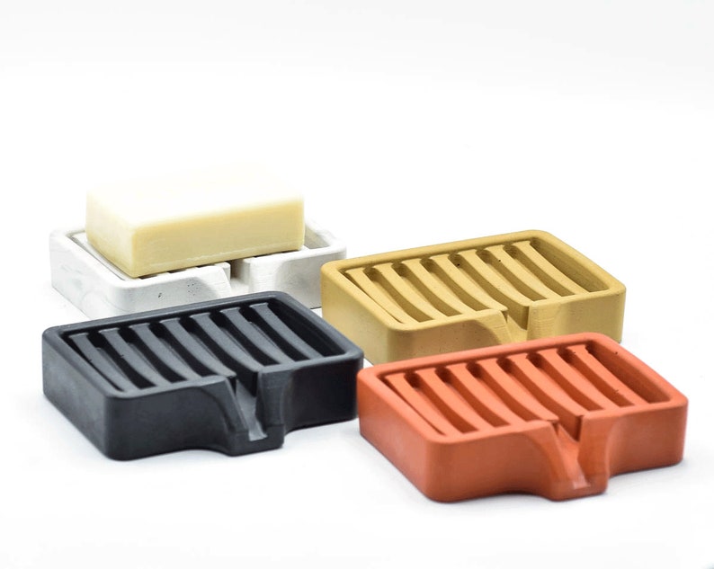 K&ouml;nnte beinhalten: Vier Seifenschalen in verschiedenen Farben: Schwarz, Wei&szlig;, Beige und Orange. Jede Schale hat eine rechteckige Form mit einem Lamellendesign und einem kleinen abgewinkelten Abschnitt zur Entw&auml;sserung. Eine Seife liegt auf der wei&szlig;en Schale.