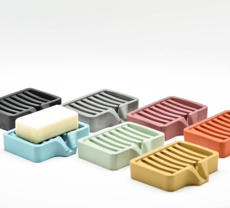 K&ouml;nnte beinhalten: Eine Sammlung farbenfroher Seifenschalen in verschiedenen Farbt&ouml;nen, darunter Schwarz, Grau, Blau, Gr&uuml;n, Rosa und Orange. Jede Schale hat eine rechteckige Form mit einem Lamellendesign und einem kleinen Ausguss. Eine Schale enth&auml;lt ein Seifenst&uuml;ck.