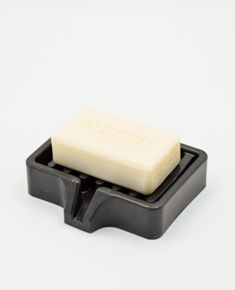 K&ouml;nnte beinhalten: Ein hellbeige Seifenst&uuml;ck mit der Aufschrift "ALL-ONE!" liegt in einer schwarzen Kunststoff-Seifenschale. Die Schale hat einen geschlitzten Boden zur Entw&auml;sserung. Der Hintergrund ist wei&szlig;.