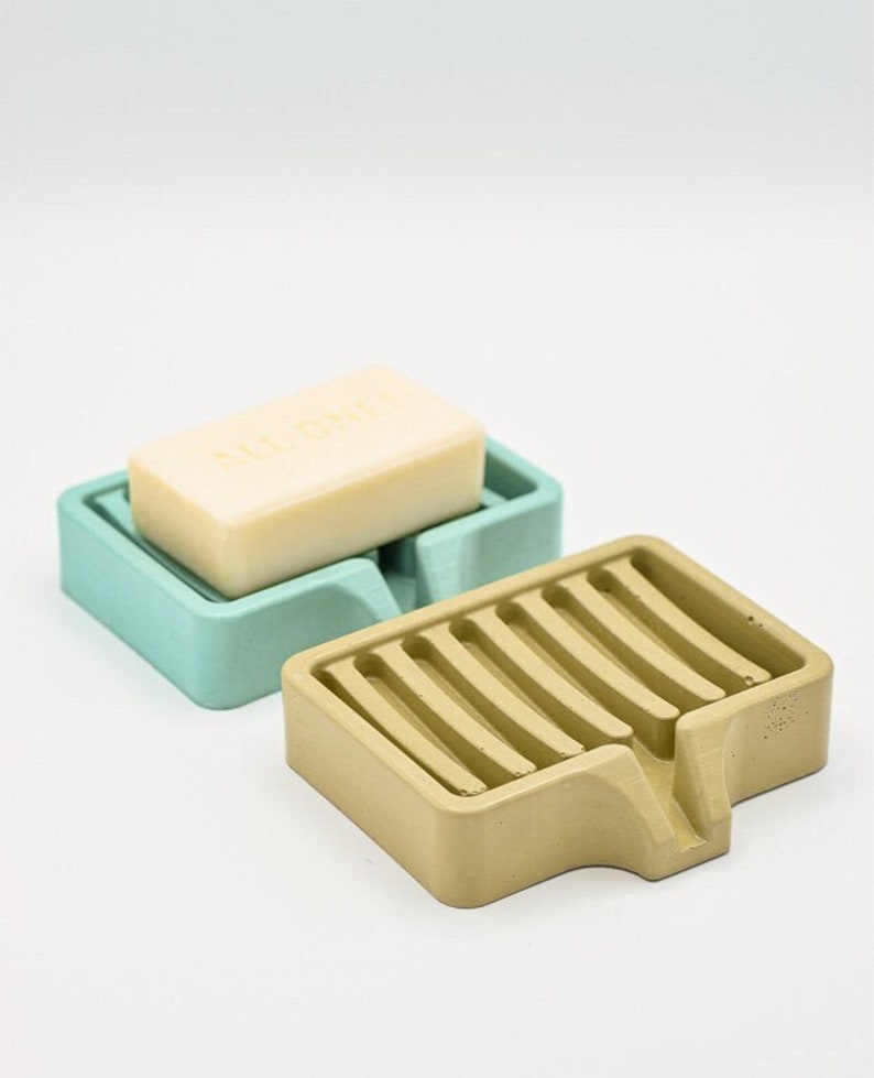 K&ouml;nnte beinhalten: Zwei Seifenschalen, eine hellt&uuml;rkis und eine beige, werden gezeigt. Die t&uuml;rkisfarbene Schale enth&auml;lt ein St&uuml;ck Seife. Die beige Schale hat ein Latten-Design. Beide sind aus einem festen Material.