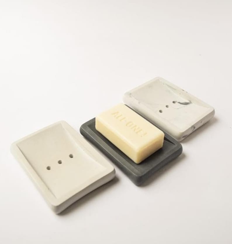 May include: Three rectangular soap dishes made of concrete in gray, white, and a marbled pattern. The dishes have drainage holes. A bar of soap is sitting on the gray dish. The soap is white and has the text "ALL ONE!" printed on it.