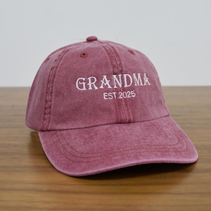 May include: A maroon baseball cap with the word "GRANDMA" embroidered in white, along with "EST 2025". The cap has a curved brim and a fabric strap closure. The hat is made of a soft, textured material.