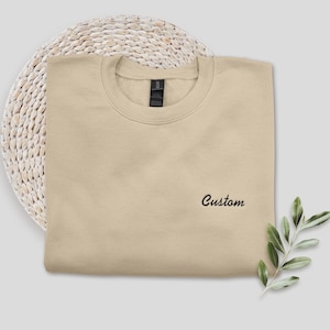 May include: A beige crewneck sweatshirt with the word "Custom" embroidered in black script. The sweatshirt is folded neatly on a woven, circular placemat. A sprig of green leaves is placed to the right of the sweatshirt.