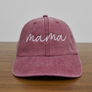 May include: A maroon baseball cap with the word "mama" embroidered in white cursive script across the front. The cap has a curved brim and a fabric strap closure on the back. The hat is made of a soft, worn material.