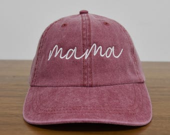 May include: A maroon baseball cap with the word "mama" embroidered in white cursive script across the front. The cap has a curved brim and a fabric strap closure on the back. The hat is made of a soft, worn material.
