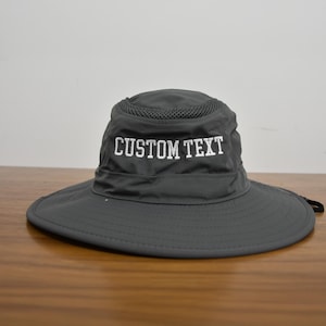 May include: A gray sun hat with a wide brim and a mesh crown. The hat features the embroidered text "CUSTOM TEXT" in white across the front. The hat is designed for sun protection and outdoor activities.