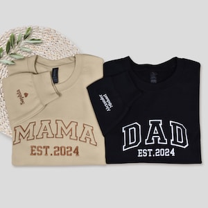 May include: Two crewneck sweatshirts, one tan and one black. The tan sweatshirt says "MAMA EST.2024" in brown lettering, with "Sophia" embroidered on the sleeve. The black sweatshirt says "DAD EST.2024" in white lettering, with "Alexander Michael" embroidered on the sleeve.