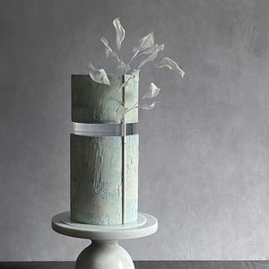 Peut inclure: Un gâteau à deux étages avec un glaçage marbré bleu clair et blanc. Le gâteau est décoré d'une seule branche blanche avec des feuilles délicates. Le gâteau est présenté sur un support à gâteau blanc.