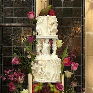 Może przedstawiać: Wielopoziomowy biały tort weselny ozdobiony figurkami cherubinów, falbanami i kwiatowymi akcentami. Ciasto jest ozdobione świeżymi winogronami, pomarańczami i gruszkami. Różowe i białe róże oraz inne kwiaty otaczają ciasto.