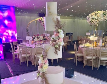 May include: A white wedding cake with gold accents and pink and white flowers. The cake is on a gold stand and is surrounded by chairs and tables.