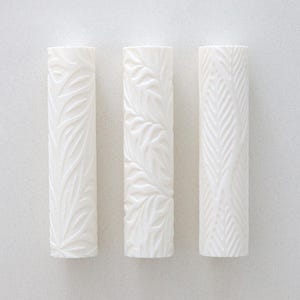 May include: Three white cylindrical objects with embossed botanical patterns. Each cylinder features a different design of leaves and foliage. The objects are arranged vertically against a plain white background.