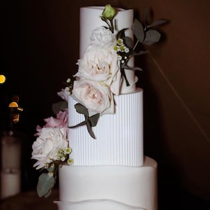 Könnte beinhalten: Eine weiße Hochzeitstorte mit vier Etagen, verziert mit weißen und rosa Rosen und Grün. Die Torte steht auf einem Holzkuchenständer.