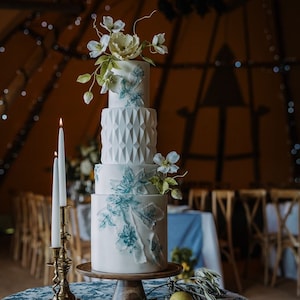 Könnte beinhalten: Eine weiße Hochzeitstorte mit vier Etagen, verziert mit blauen Blumenmustern und weißen Blüten. Die Torte steht auf einem Holzkuchenständer und ist von Grünpflanzen und Kerzen umgeben.