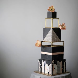 May include: A black and gold wedding cake with geometric designs and peach-coloured flowers. The cake is made of multiple tiers and is decorated with a geometric pattern.