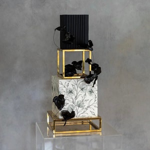 Peut inclure: Un gâteau de mariage à trois étages avec des motifs floraux noirs et blancs. L'étage inférieur est blanc avec des motifs floraux noirs, l'étage du milieu est noir et l'étage supérieur est un gâteau carré noir. Le gâteau est décoré d'accents floraux noirs et repose sur un support doré.