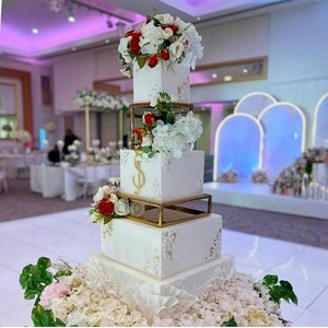 Peut inclure: Un gâteau de mariage à quatre étages décoré de fondant blanc et d'accents dorés. Le gâteau est orné de fleurs blanches et rouges, et le niveau supérieur porte une lettre dorée 'S'.