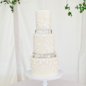Puede incluir: Un pastel de boda blanco de cuatro pisos decorado con delicados diseños florales blancos. El pastel está sobre un soporte para pastel blanco.