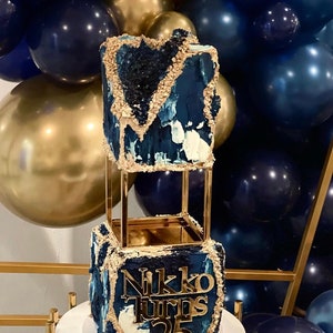 Peut inclure: Un gâteau à deux étages décoré de glaçage bleu et or. Le gâteau est sur un support doré et porte une pancarte dorée qui dit "Nikko Turns 25".