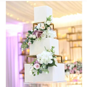 May include: A four-tier white square wedding cake with gold geometric tiers and floral decorations. The flowers are white, pink, and purple.