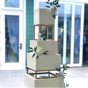 Peut inclure: Un gâteau de mariage blanc carré à quatre étages avec des supports de gâteau géométriques dorés et des fleurs blanches.