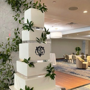 Może przedstawiać: Biały tort ślubny z czterema piętrami, ozdobiony zielenią i czarnym monogramem. Tort stoi na przezroczystym stojaku akrylowym.