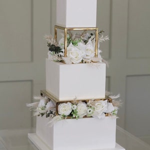 Peut inclure: Un gâteau de mariage blanc à quatre étages avec des supports de gâteau géométriques dorés et des fleurs blanches.