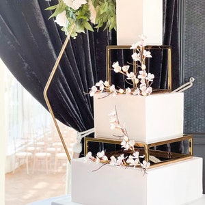 Peut inclure: Un gâteau de mariage blanc à quatre étages, chaque étage est une boîte carrée avec un cadre doré. Le gâteau est décoré de fleurs blanches et de verdure.