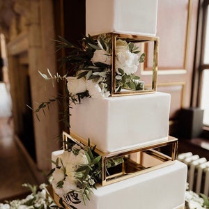 Peut inclure: Un gâteau de mariage carré blanc avec des étages géométriques dorés. Le gâteau est décoré de fleurs blanches et de verdure.