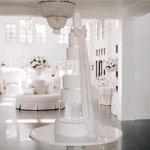 Puede incluir: Un pastel de boda blanco con niveles de acrílico transparente y una cinta de satén blanco. El pastel está sobre una mesa redonda blanca con una base de pedestal blanco.