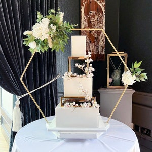 Peut inclure: Un gâteau de mariage blanc avec de délicates fleurs roses sur chaque étage, présenté sur un support à gâteau géométrique doré. Le gâteau est décoré d'un arrangement floral de fleurs blanches et de verdure.