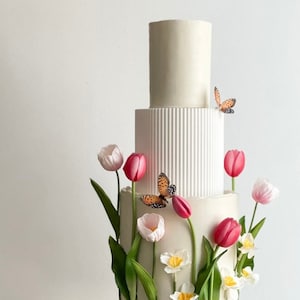 Könnte beinhalten: Eine weiße Hochzeitstorte, verziert mit rosa und weißen Tulpen, grünen Blättern und orangefarbenen Schmetterlingen. Die Torte steht auf einem weißen Tortenständer mit einem Holzsockel.