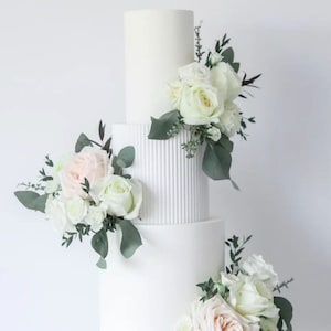 Könnte beinhalten: Eine dreistöckige weiße Hochzeitstorte, verziert mit weißen und rosafarbenen Rosen und Grün. Die Torte steht auf einem weißen Tortenständer. "Sally Cooper Cake Artist" steht in der rechten unteren Ecke des Bildes.