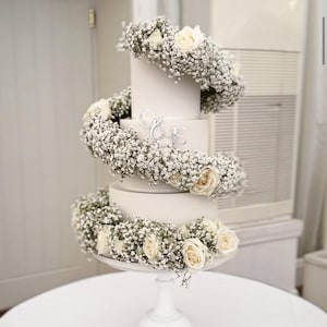 Puede incluir: Una tarta de boda blanca de tres pisos decorada con rosas blancas y paniculata. La tarta está sobre un pedestal blanco y presenta iniciales plateadas. Las flores están dispuestas en espiral alrededor de la tarta.