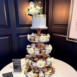 Pode incluir: Um bolo de casamento branco com detalhes em folha de ouro fica em cima de um suporte de cupcake de quatro andares. O suporte é decorado com flores brancas e contém uma variedade de cupcakes, incluindo red velvet, chocolate e caramelo com sal marinho, merengue de limão e laranja vegana.