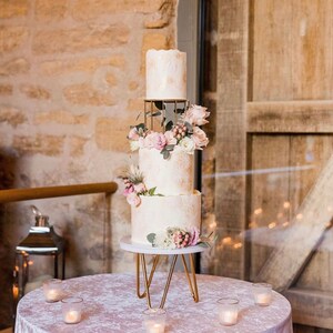 Può includere: Una torta nuziale a tre piani con glassa bianca e delicati disegni floreali. La torta è decorata con fiori rosa e bianchi e verde, e si trova su un supporto in metallo dorato.