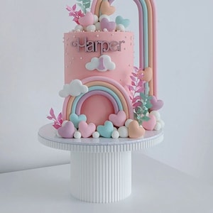 Könnte beinhalten: Ein rosa Kuchen, verziert mit einem Regenbogen, Wolken und Herzen. Der Kuchen steht auf einem weißen Kuchenständer und trägt vorne die Aufschrift "Harper 1".
